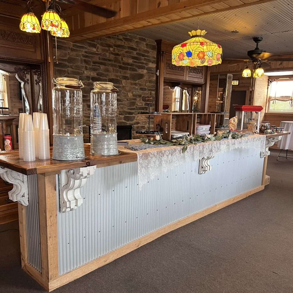 A rustic refreshment bar featuring glass water dispensers, lace table runners, and decorative lanterns with candles, set in a warm wooden interior with stained-glass lighting in Alliance, Ohio.